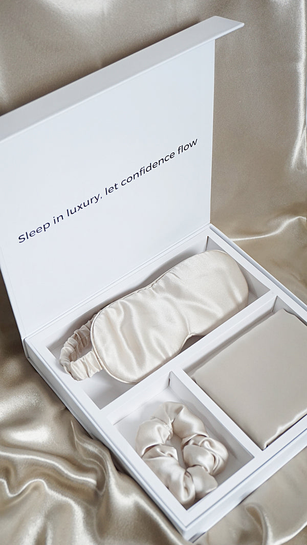 Silk sleep mask and scrunchie set in a white box with 'Sleep in luxury, let confidence flow' text on a satin background.