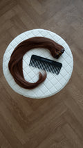 Brown hair extensions with a comb on a white cushioned stool.