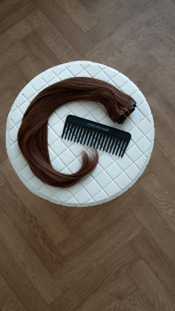 Brown hair extensions with a comb on a white cushioned stool.